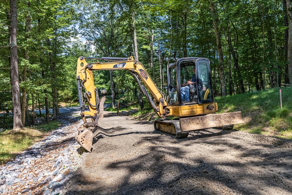 Excavation services in progress using a compact excavator to grade and level a gravel access road on a commercial construction site in a wooded area.