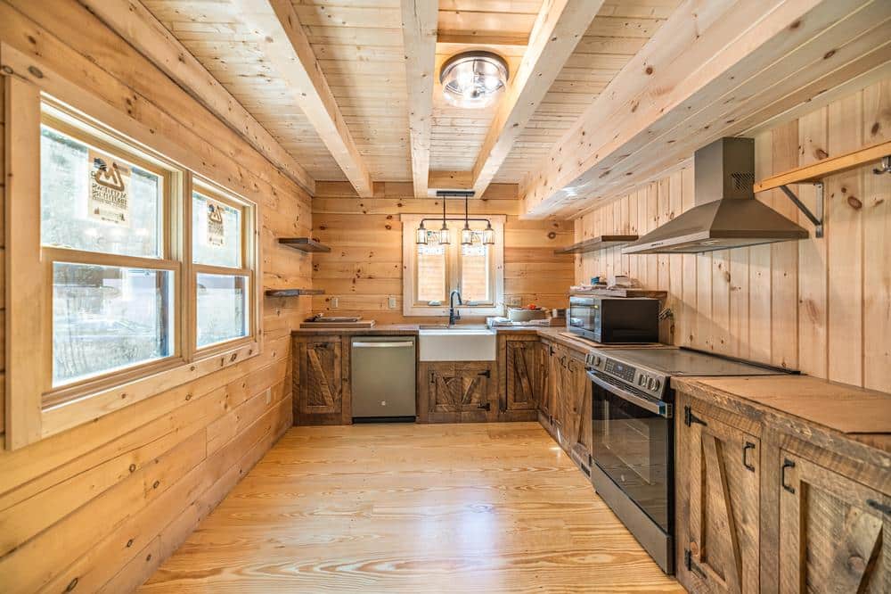 Residential construction kitchen renovation featuring a rustic log-style interior with custom wood cabinetry, farmhouse sink, stainless steel appliances, exposed beam ceiling, and natural light from multiple windows.
