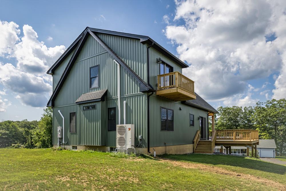 Residential construction exterior showing a newly built custom home with vertical siding, an upper-level balcony, rear deck, and landscaped hillside, highlighting a completed residential build in a rural setting.