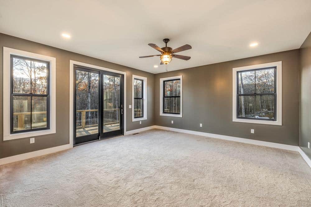 Residential construction interior showing a finished bedroom with neutral walls, plush carpet flooring, ceiling fan, and large windows providing natural light and wooded views.