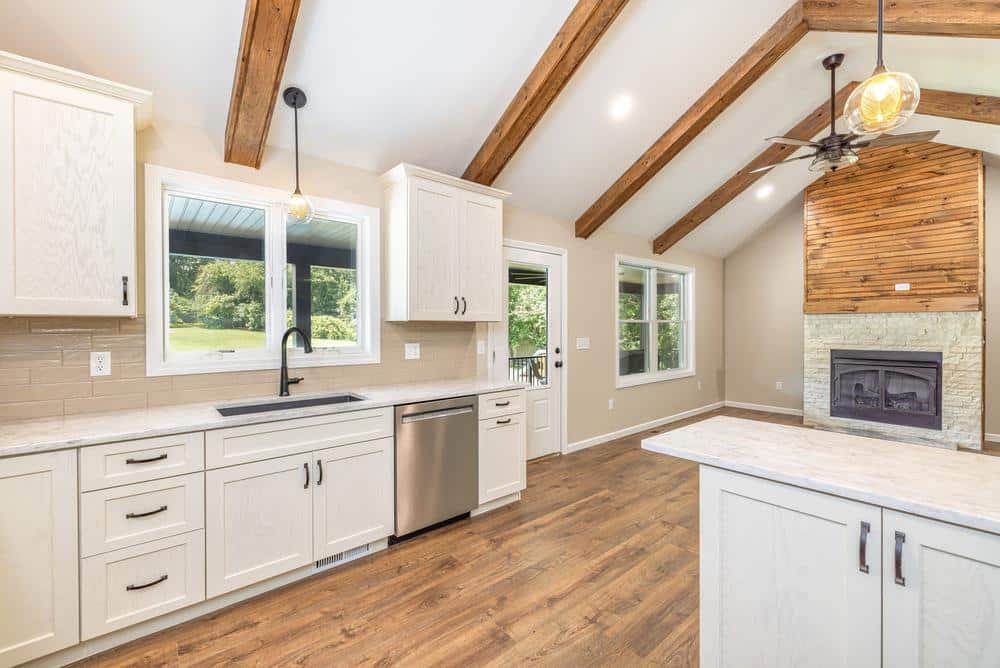 Residential construction interior featuring an open-concept kitchen renovation with white cabinetry, stone countertops, exposed wood beams, and a connected living area with fireplace.