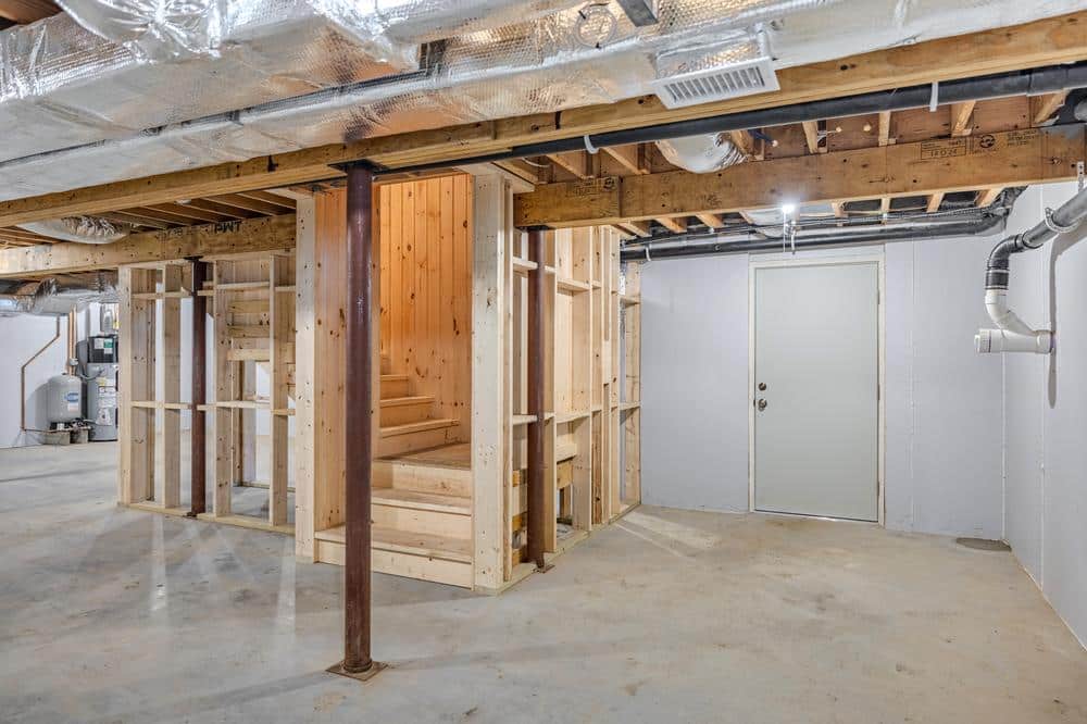Residential construction interior showing an unfinished basement with framed stairwell, exposed wood framing, HVAC ductwork, utility systems, and concrete flooring ready for future build-out.