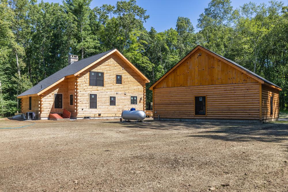 Residential construction exterior showing a completed log-style home and detached garage on a cleared, wooded property with finished grading and site work.