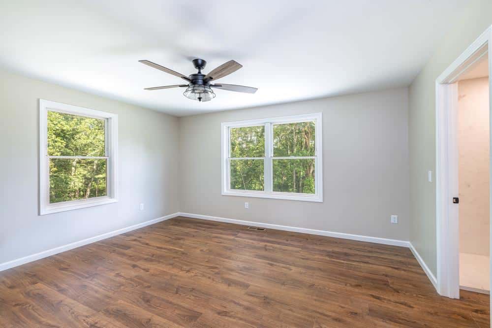 Residential construction interior featuring a finished bedroom with wood-look flooring, ceiling fan, neutral wall colors, and large windows providing natural light in a newly built home.