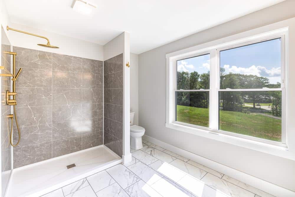 Modern walk-in shower with large gray tile walls, gold rain shower fixtures, marble-look tile flooring, and oversized window bringing in natural light, showcasing custom residential bathroom construction.