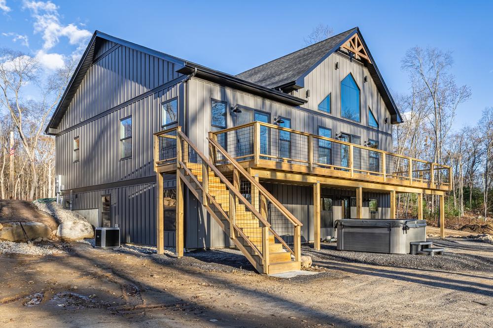Residential construction exterior showing a custom-built home with vertical metal siding, an elevated deck with stairs, and finished site work surrounding the foundation.