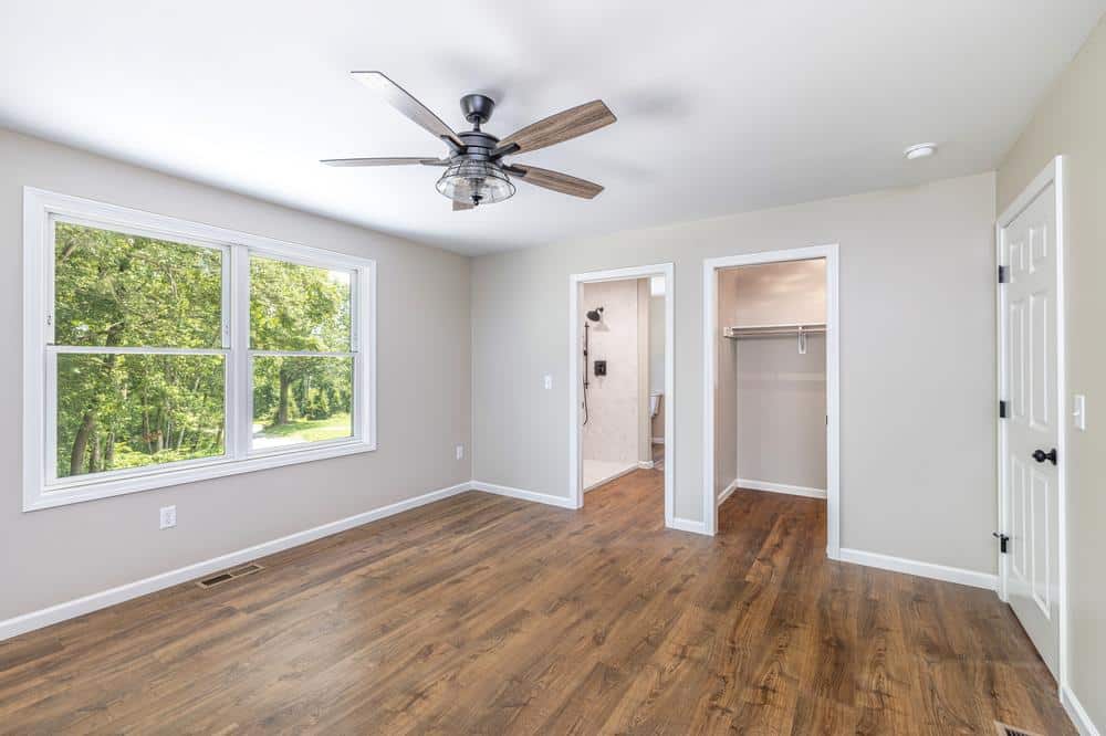 Residential construction interior showing a finished bedroom with wood-look flooring, ceiling fan, neutral walls, and an adjoining closet and bathroom entry in a newly built home.