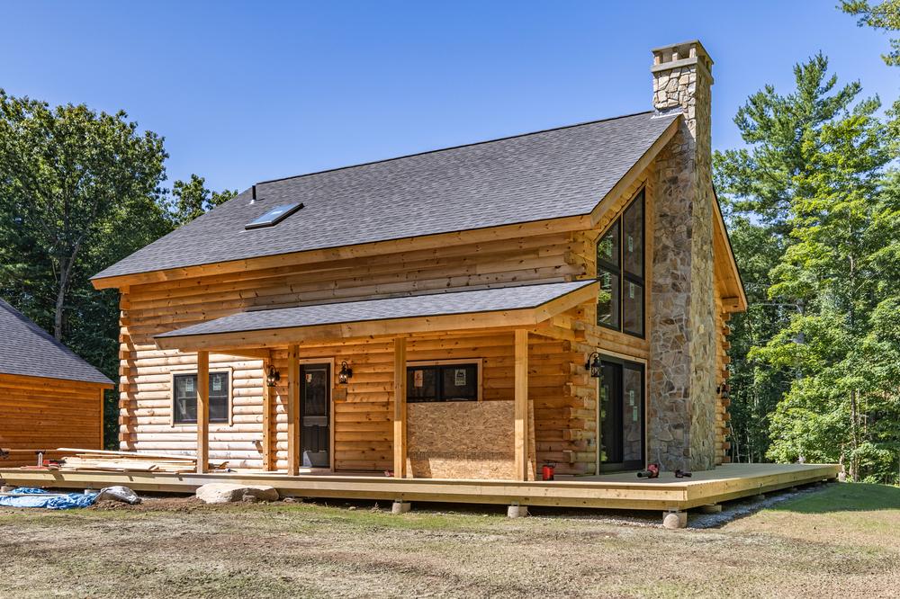 Residential construction exterior showing a log-style home under construction with a stone chimney, covered porch, wood siding, and wraparound deck being completed on a wooded property.