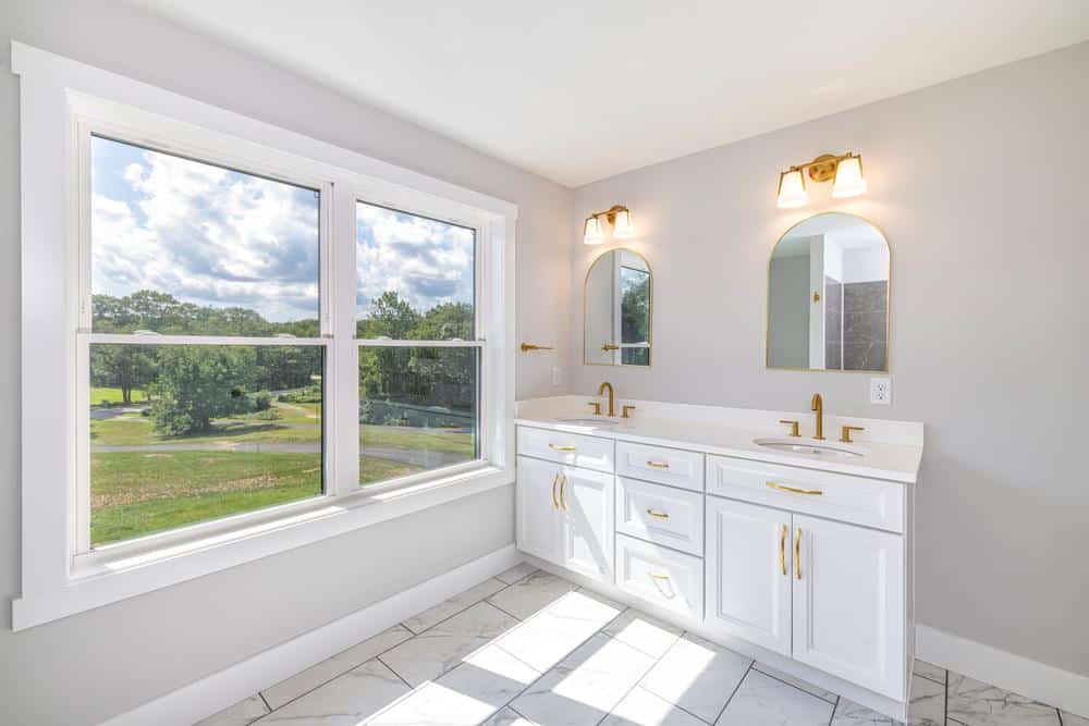 Bright custom bathroom with double vanity featuring white cabinetry, gold fixtures, arched mirrors, tiled floor, and large window overlooking a landscaped outdoor view, highlighting high-end residential construction.