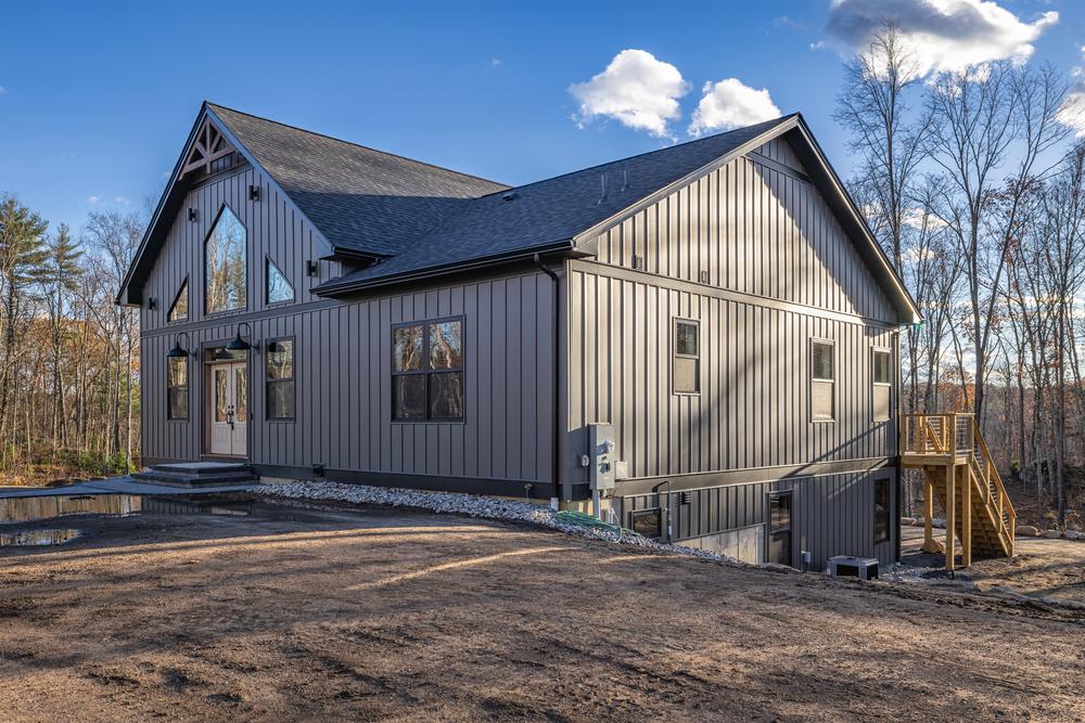 Residential construction exterior showing a newly built home with vertical metal siding, elevated deck and stairs, and completed foundation and site work on a wooded property.