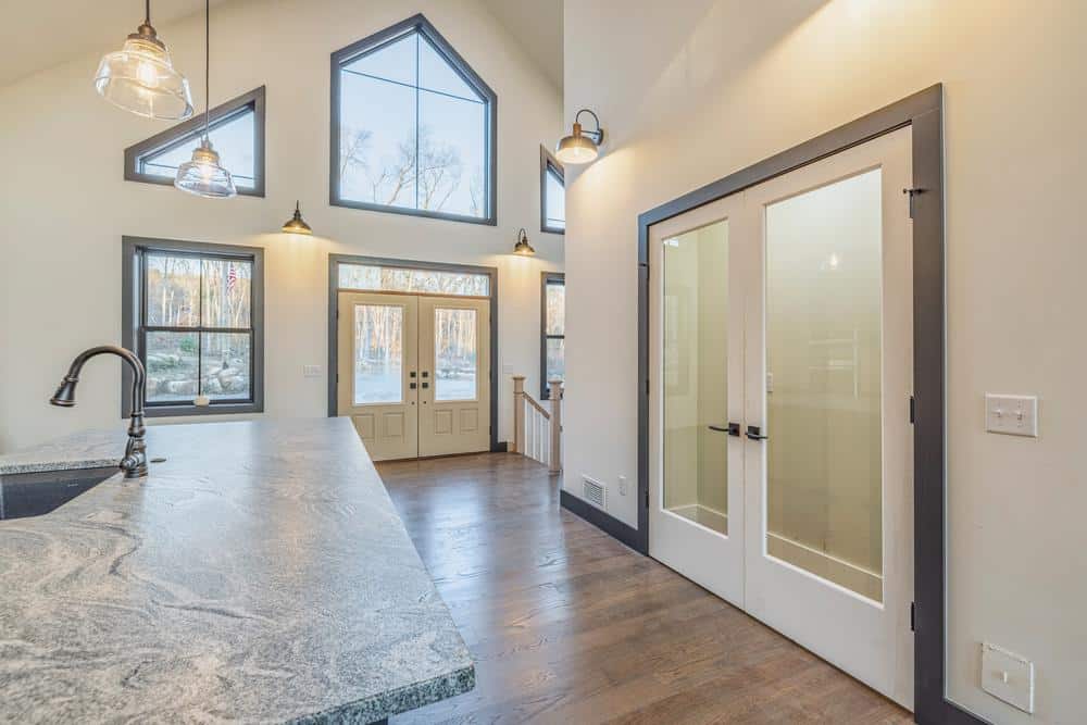 Residential construction interior showing a completed kitchen renovation with stone countertops, modern lighting, vaulted ceilings, and large windows bringing in natural light.