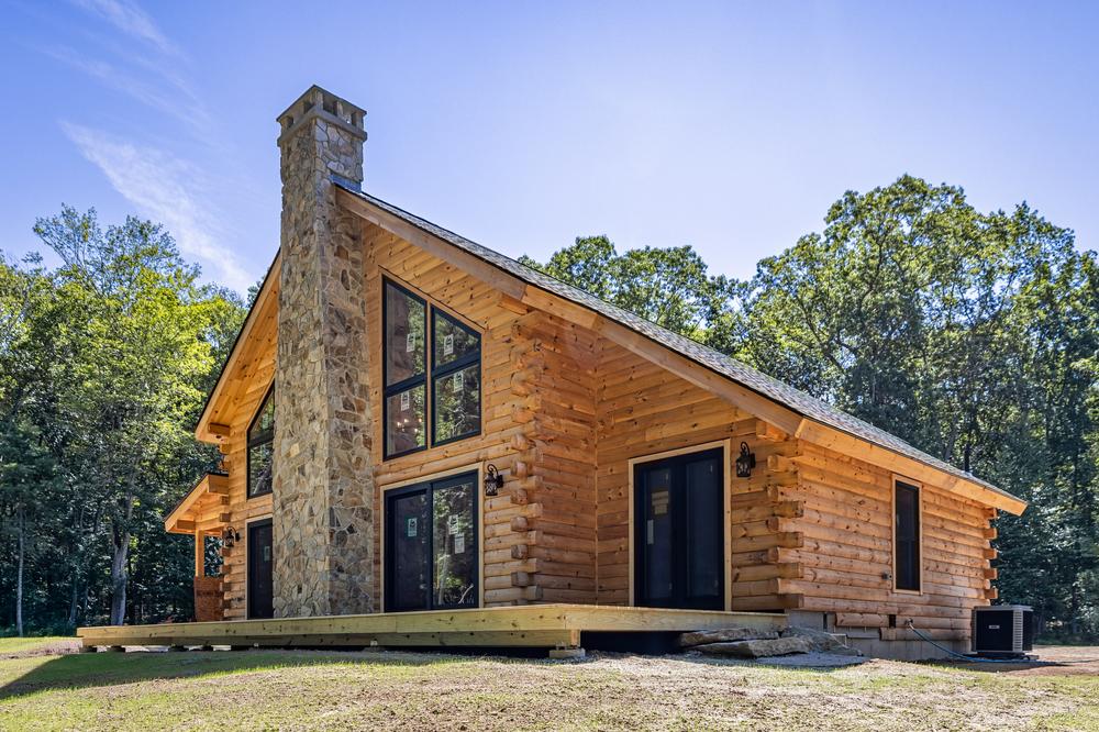 Residential construction exterior showcasing a newly completed log-style home with a stone chimney, wood siding, large windows, and a wraparound deck set on a wooded property.