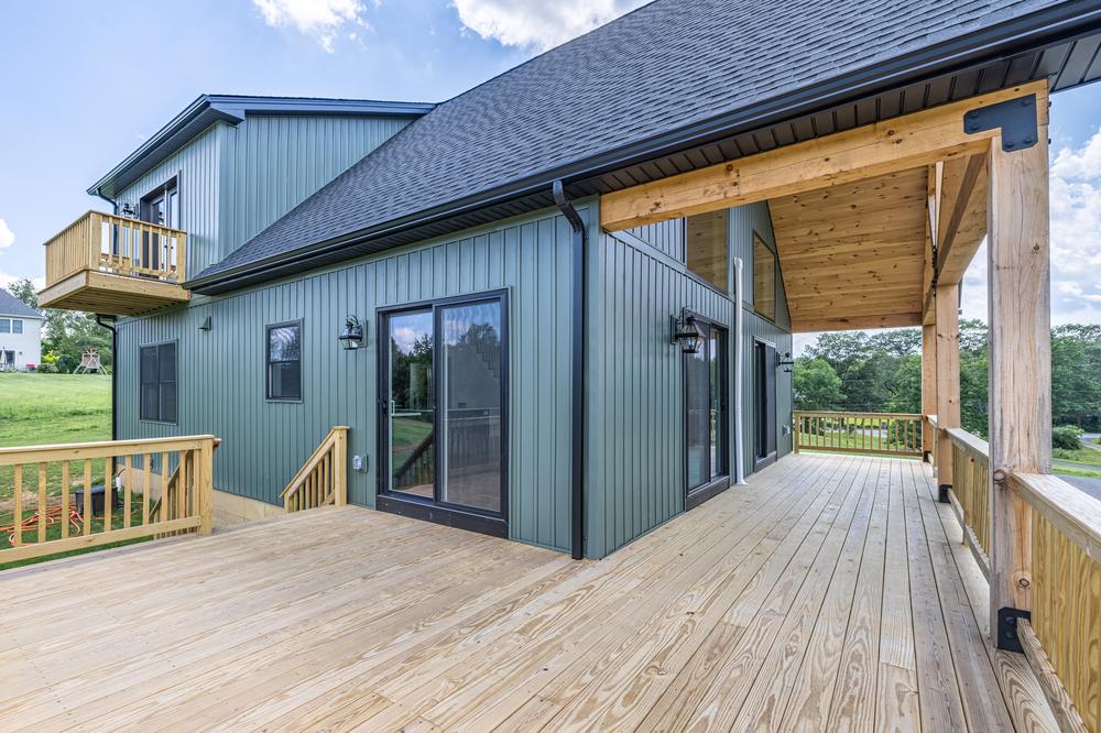 Custom residential construction featuring a large wraparound wooden deck with covered porch, sliding glass doors, and green vertical siding, showcasing outdoor living space and quality home building craftsmanship.