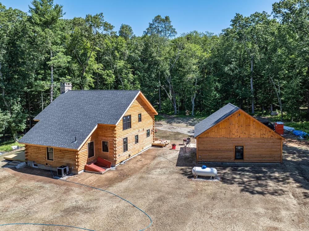 Residential construction project showing a newly built log-style home and detached garage on a wooded property with site work nearing completion.